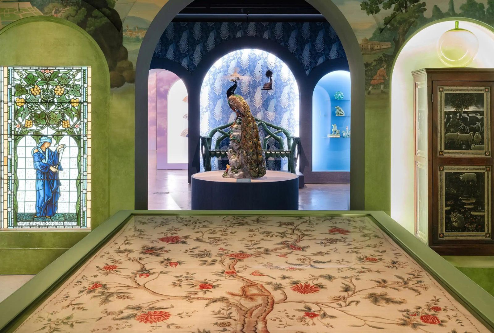 A green room with arches, on the left there is a stained glass window, on the right is a lit up wooden capinet. In the centre is a large sculpture of a male peacock and in front of it is a large rug with a flowering tree.