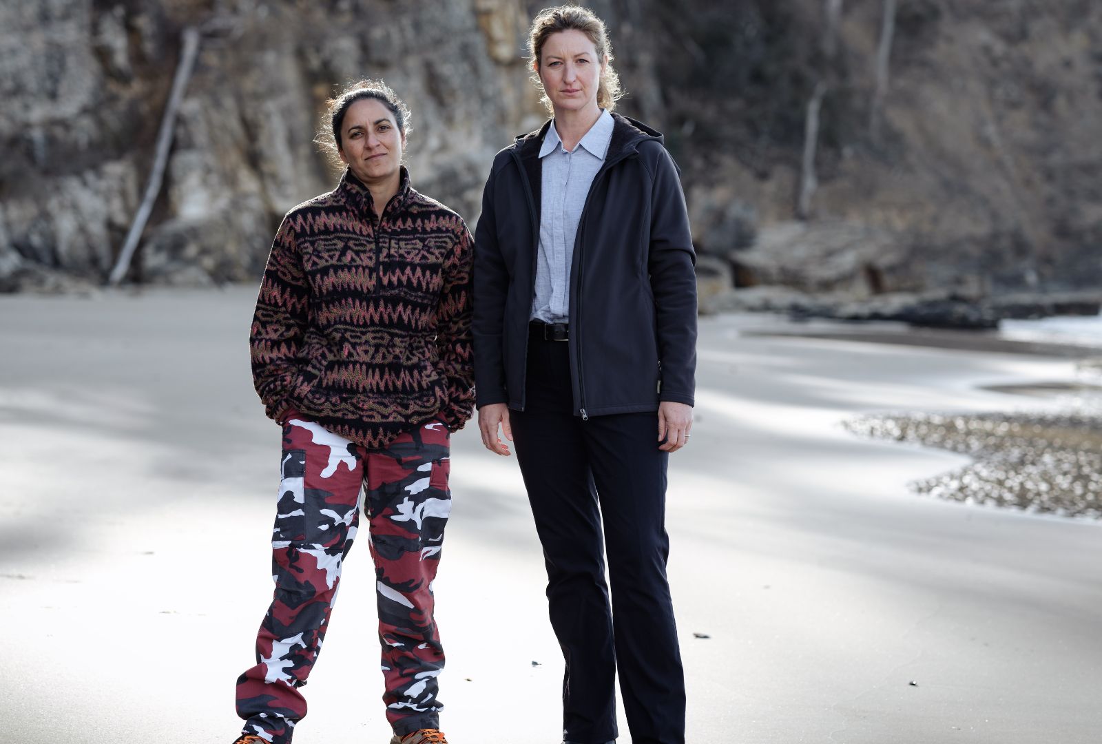 Madeleine Sami as Eddie Redcliffe and Kate Box (Acting, 2003) as Dulcie Collins in Amazon Prime's 'Deadloch', They stand on a beach in winter clothing looking towards the camera.