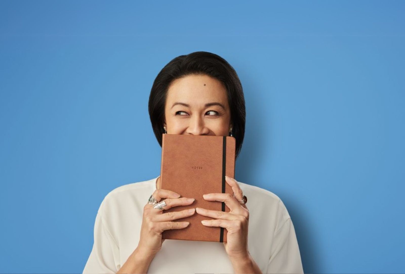 A woman stands in front of a blue background holding a brown book over her mouth.