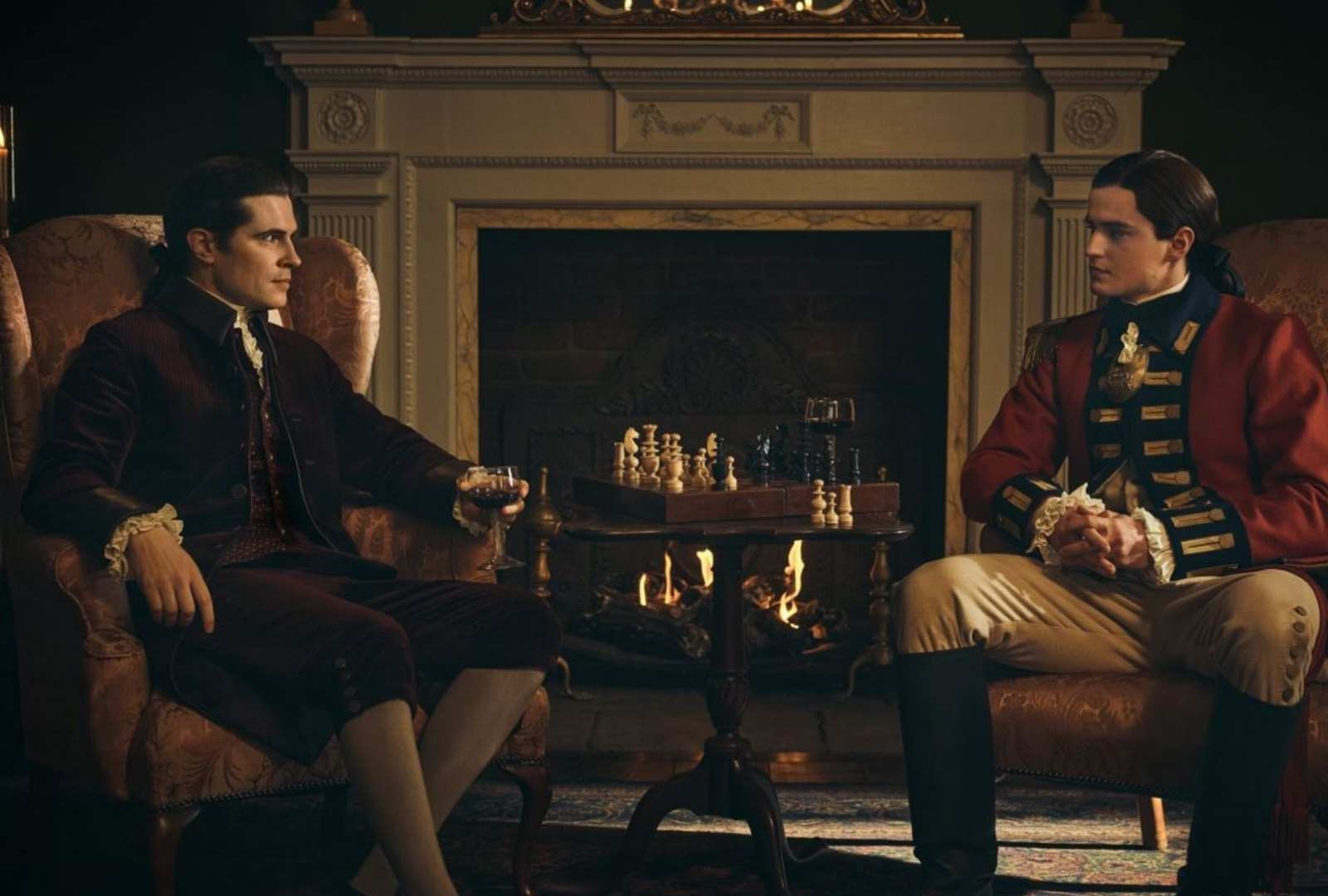 From left to right: David Berry as Lord John Grey and Charles Vandervaart as William Ransom in Binge's 'Outlander'. They sit across from each other in a lounge room playing chess while drinking wine.