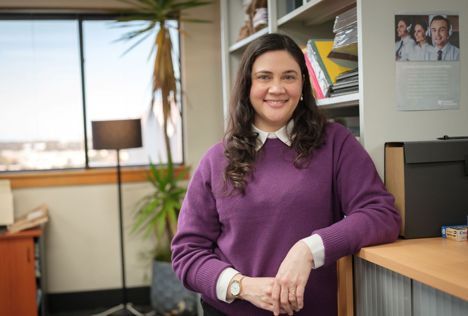 Shari Sebbens as Greta King in 'The Office'. Shari is leaning on a desk in an office building smiling with her hands in front of her.