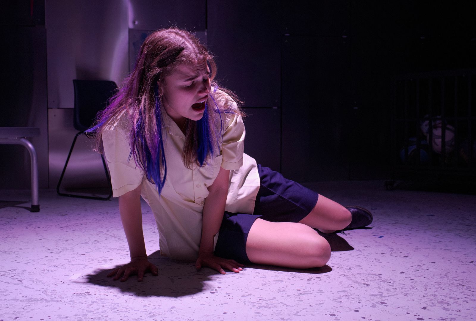 Clare Hughes performing as Sam in 'Sitting Screaming'. She is on the floor in a school uniform with blue streaks in her hair, screaming.