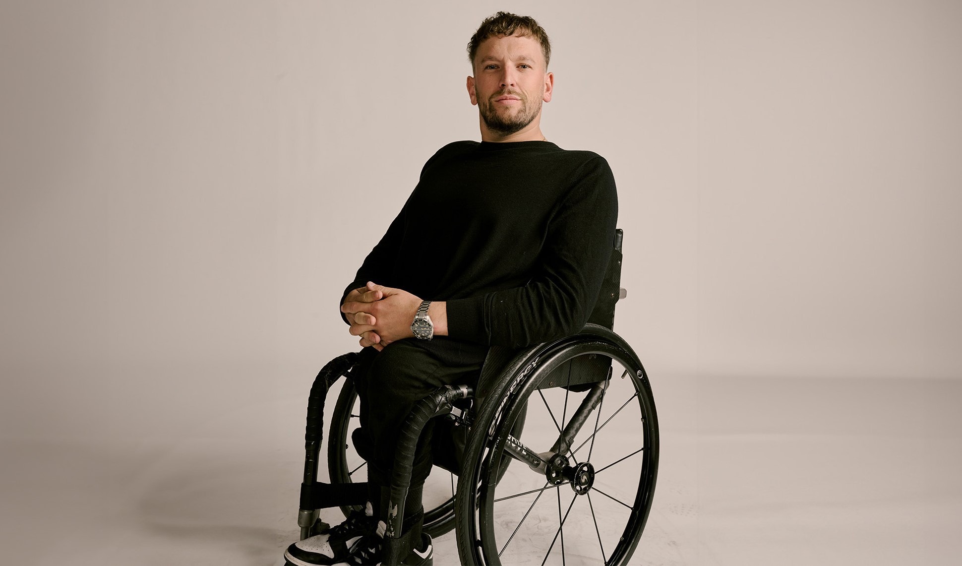 Dylan Alcott posed in front of a white background wearing a black, long-sleeved shirt and sitting in a wheelchair.